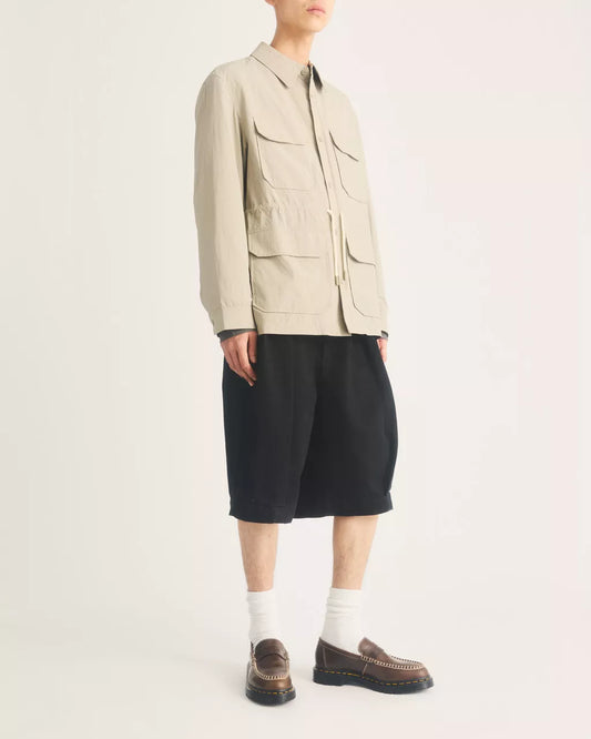 A model wearing a light beige utility jacket with multiple front pockets, black shorts, white socks, and brown leather loafers, photographed against a clean off-white studio background.