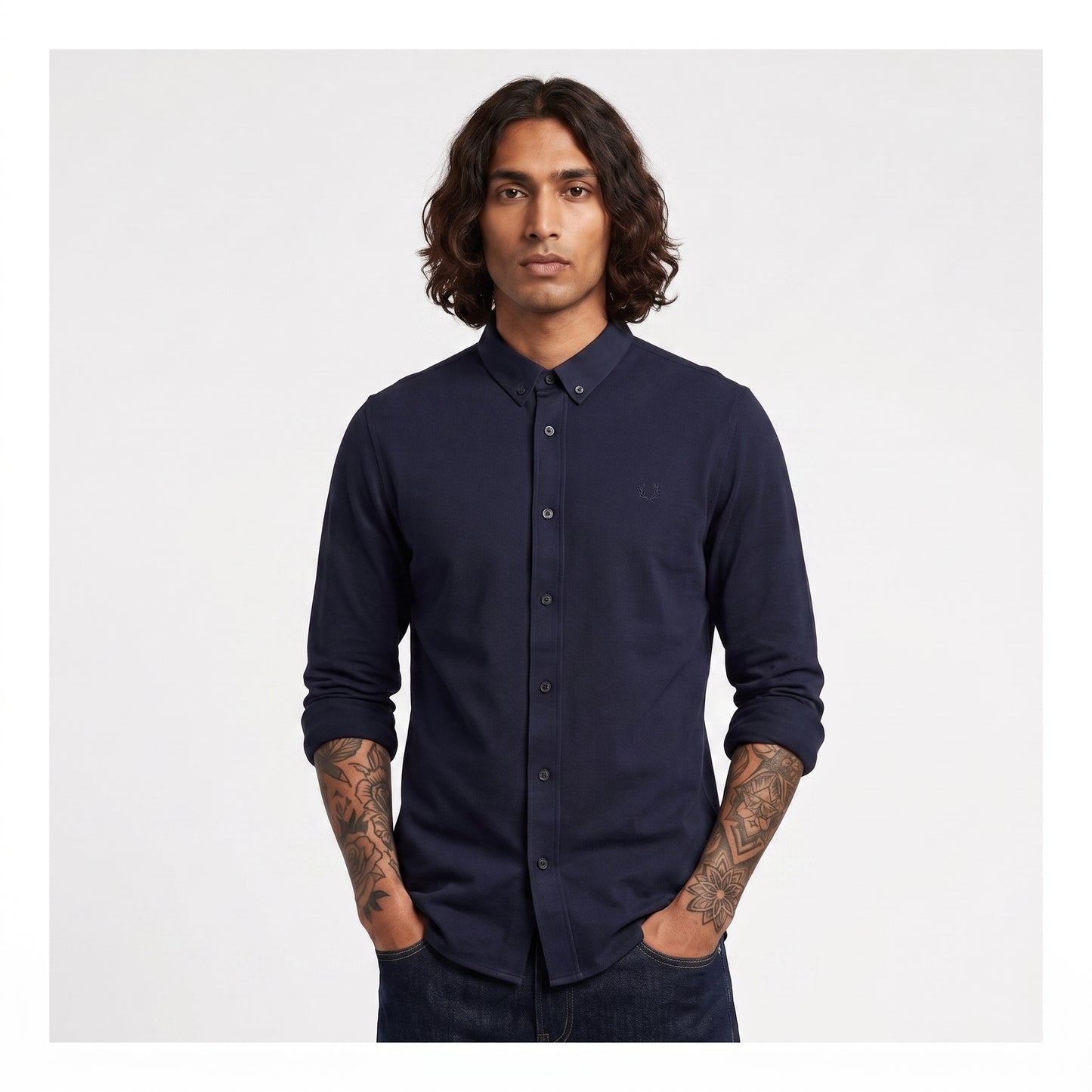 A man with long dark hair and tattoos wearing a dark navy long-sleeve button-down shirt, standing against a light gray background.