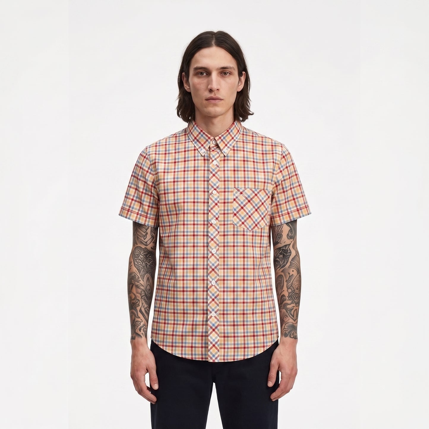 A man with long dark hair and tattoos stands against a plain white background wearing a short-sleeve button-up shirt with a red, orange, and blue plaid pattern.