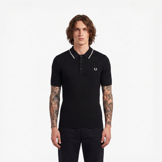 A man wearing a black ribbed short-sleeve polo shirt with white piping on the collar and a small embroidered laurel wreath logo on the chest, photographed against a plain white studio background.