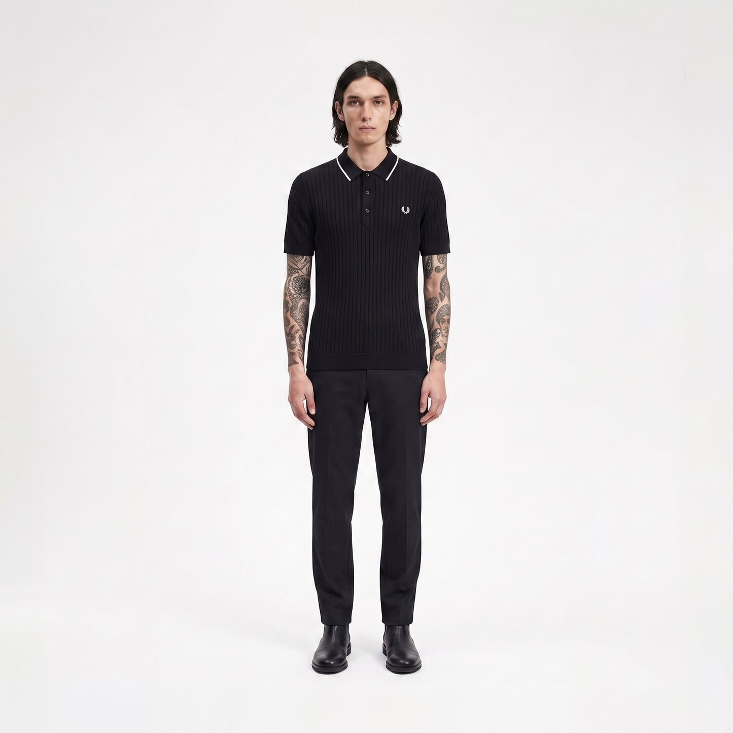 A man stands facing forward in a black ribbed short-sleeve polo shirt with white piping and a small embroidered laurel wreath logo on the chest, paired with black trousers and black shoes, against a plain light gray studio background.
