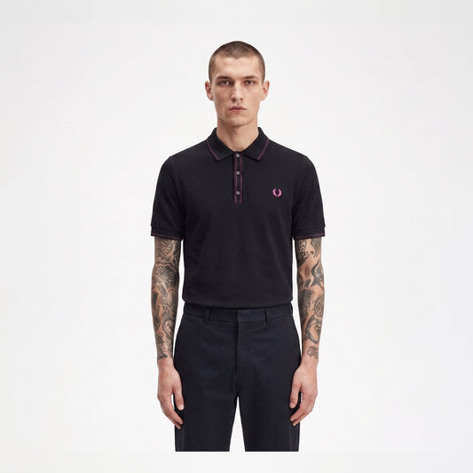 A man modeling a black short-sleeve polo shirt with a dark red collar and placket, featuring the signature pink and white embroidered laurel branch logo on the left chest, paired with dark trousers against a plain white background.