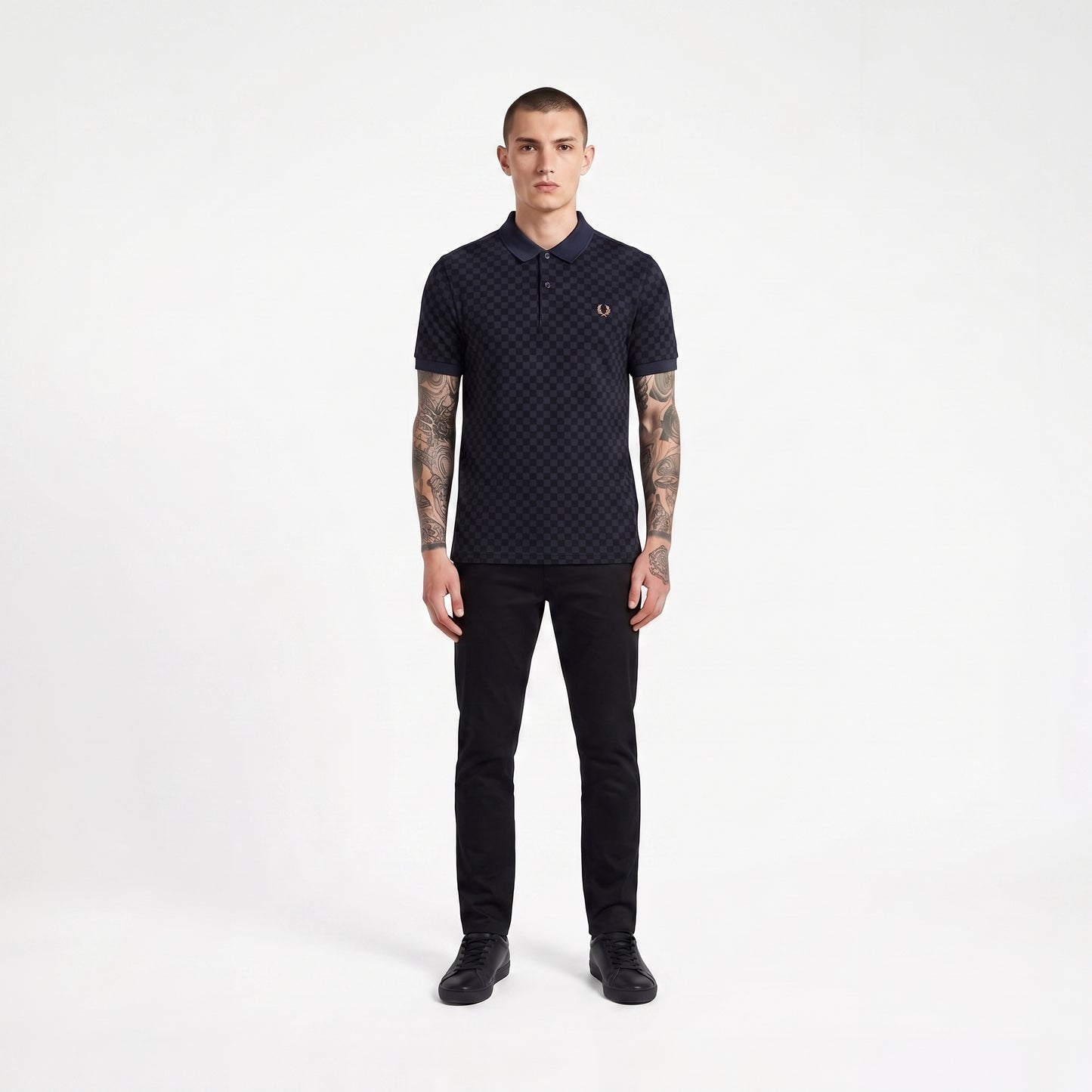 A man modeling a dark navy short-sleeve polo shirt with a subtle geometric pattern and a small embroidered laurel wreath logo on the chest, paired with black trousers and black shoes, against a plain white studio background.