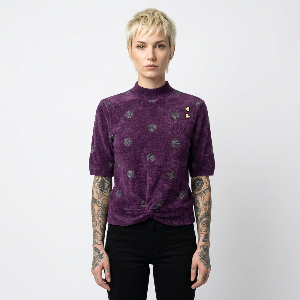A model with short blonde hair and tattooed arms wearing a purple short-sleeve mock neck top with a twisted hem and small gold heart embellishments, paired with black pants, against a plain white background.