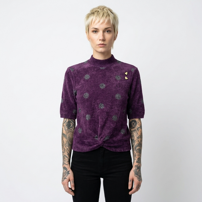 A model with short blonde hair and tattooed arms wearing a purple short-sleeve mock neck top with a twisted hem and small gold heart embellishments, paired with black pants, against a plain white background.