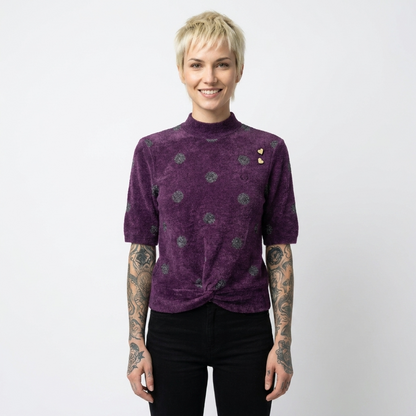 A model with short blonde hair and tattoos wearing a purple short-sleeve mock neck top with a textured pattern, paired with black pants, against a plain light gray background.