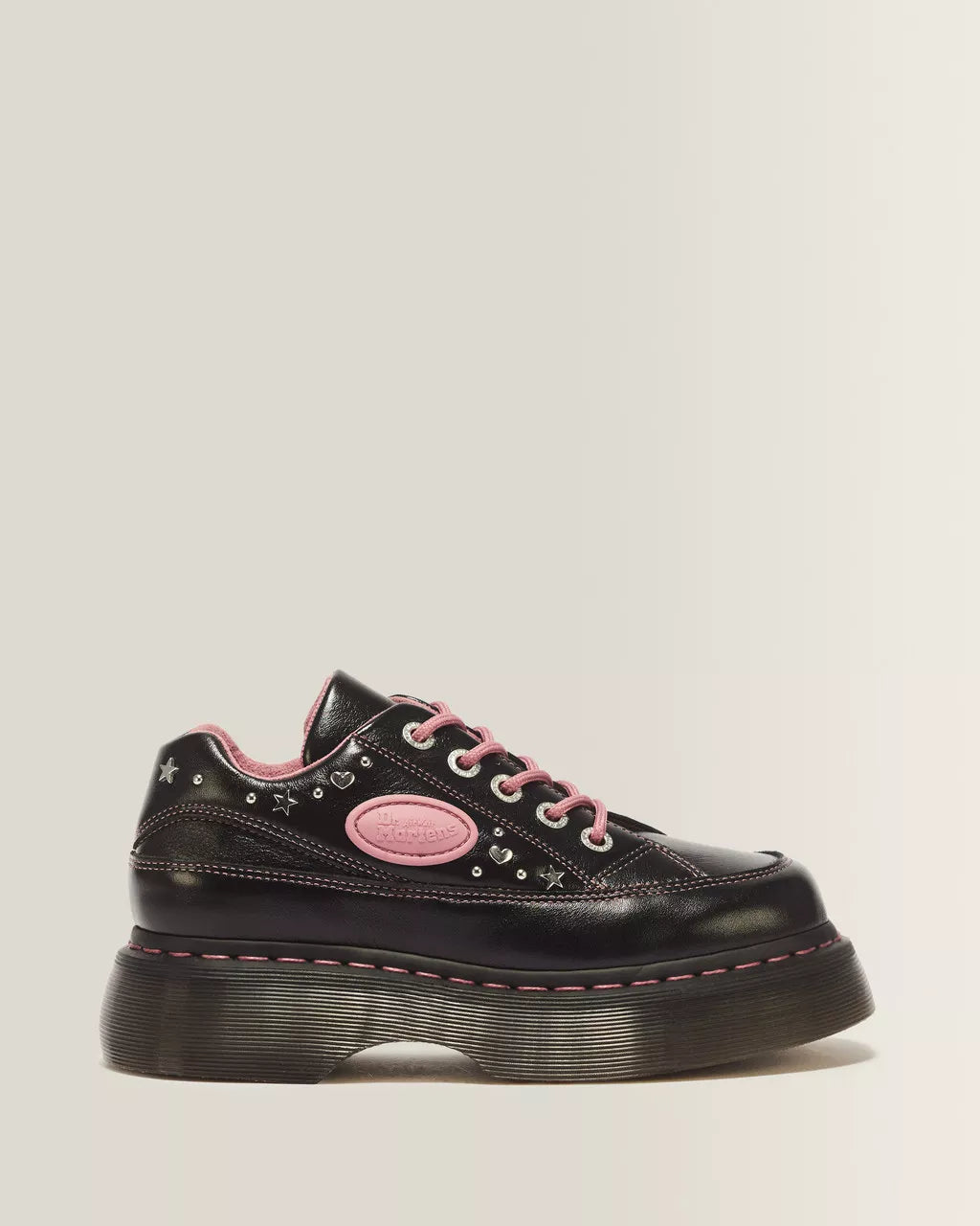 A black leather platform sneaker with pink laces, a pink oval patch, and pink stitching, decorated with silver studs and star details, shown against a light beige background.