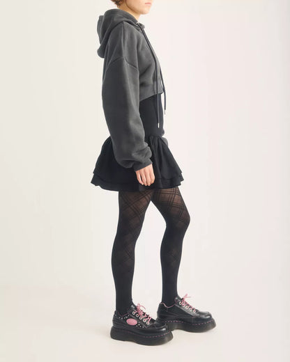 A model shown from the side wearing a dark gray and black color-blocked hoodie dress, black fishnet tights, and black platform shoes with pink laces against a plain off-white background.