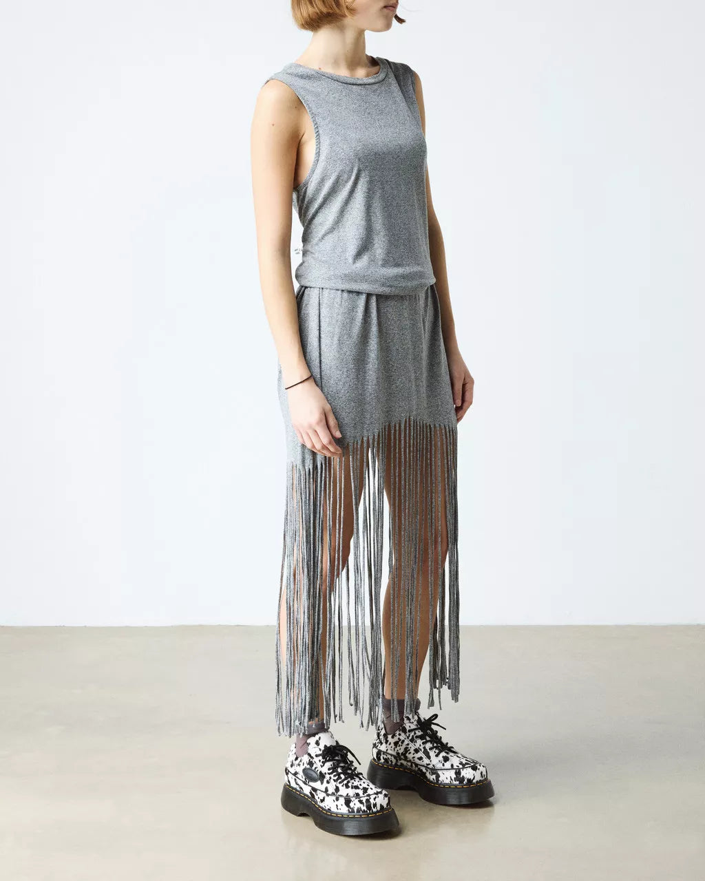 Person wearing a gray sleeveless top and fringe skirt with black and white shoes on a plain background