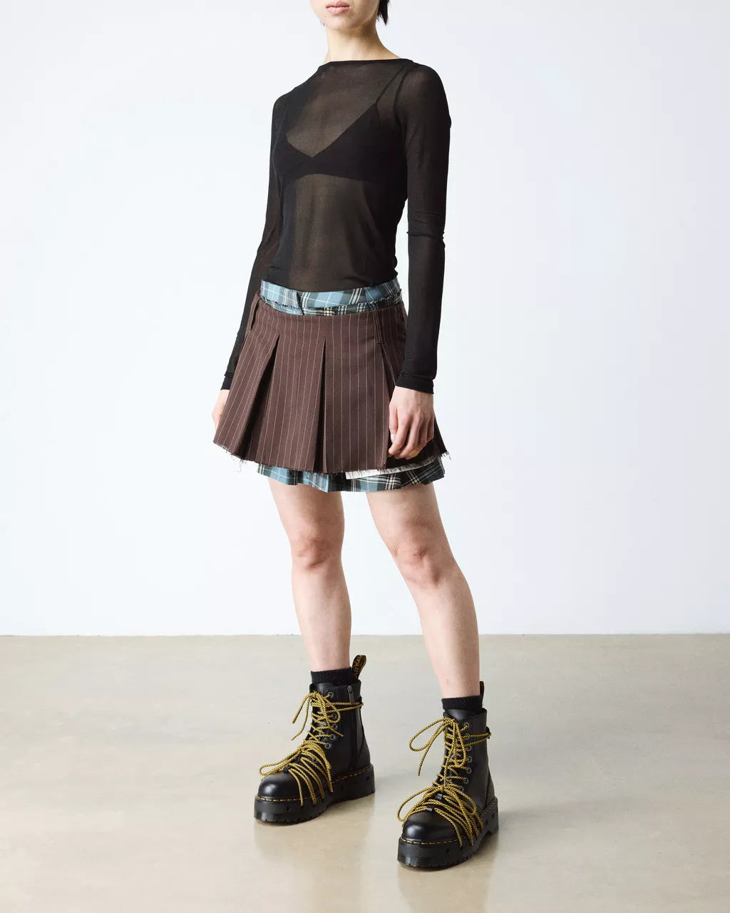 Person wearing a black sheer top, plaid skirt, and black boots on a plain background