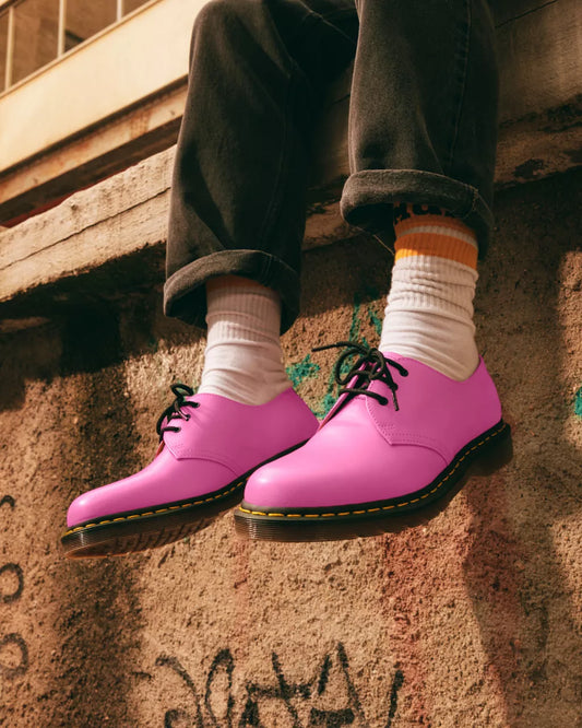A person wearing smooth leather oxford shoes in thrift pink color, with the sole of the shoes facing forward, resting their feet on a wall with graffiti.