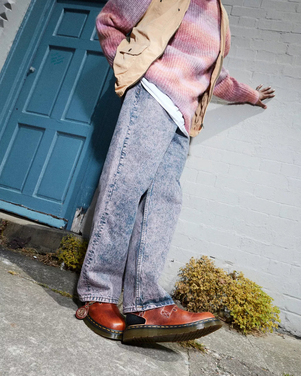 Person wearing a pink sweater, beige jacket, blue jeans, and brown shoes leaning against a white wall with a blue door.