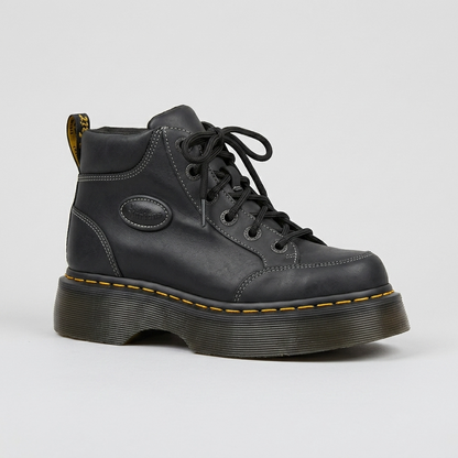 A Dr Martens Buzz 6i black leather lace-up ankle boot with a chunky black platform sole, yellow contrast stitching, and a gold pull tab, shown on a light gray background.