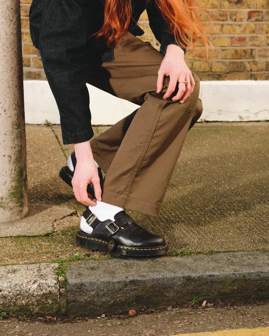 Person wearing brown pants and black Dr. Martens shoes on a sidewalk.