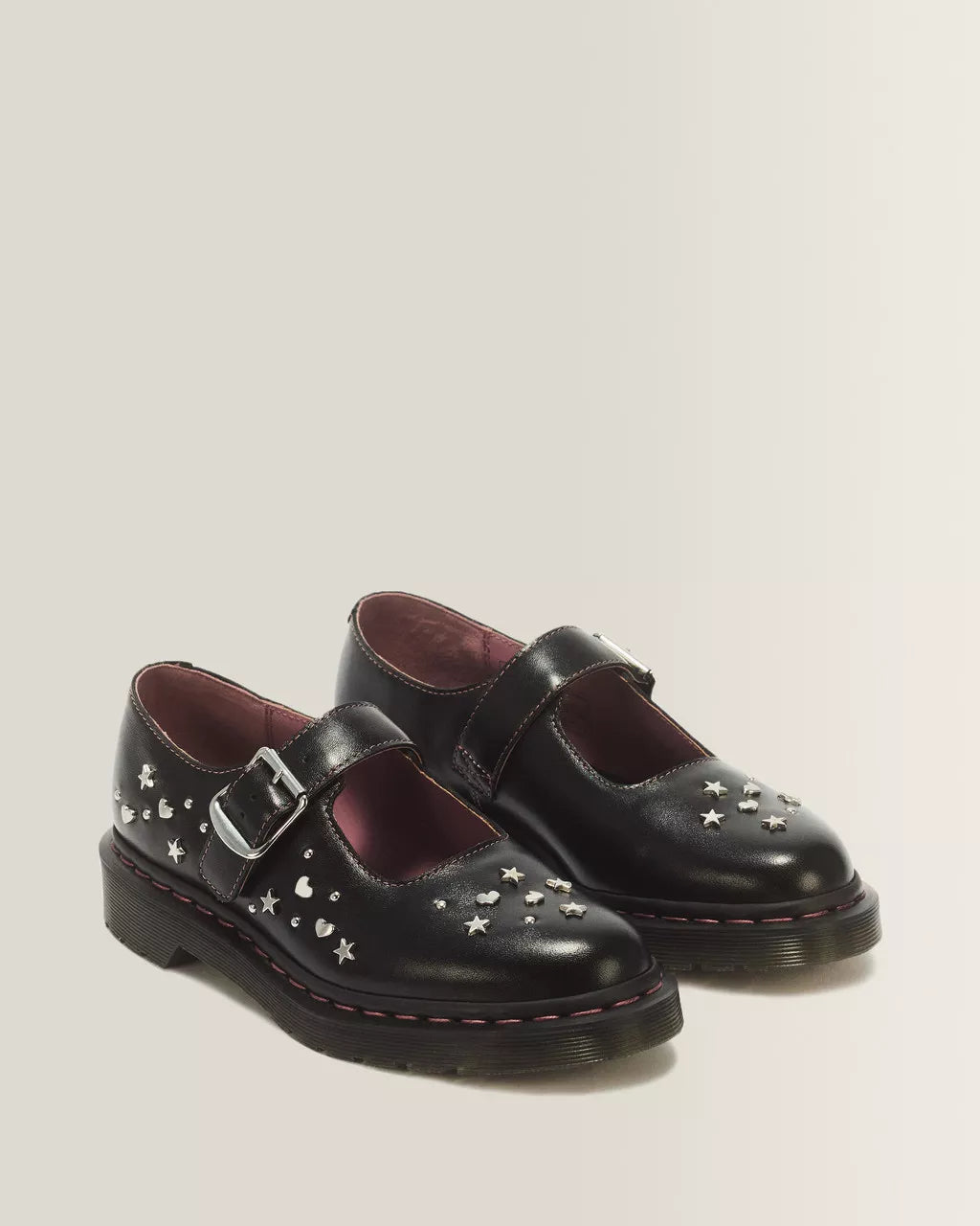A pair of black leather Mary Jane shoes decorated with silver star and heart studs, featuring a buckle strap and a thick black sole, set against a plain light beige background.