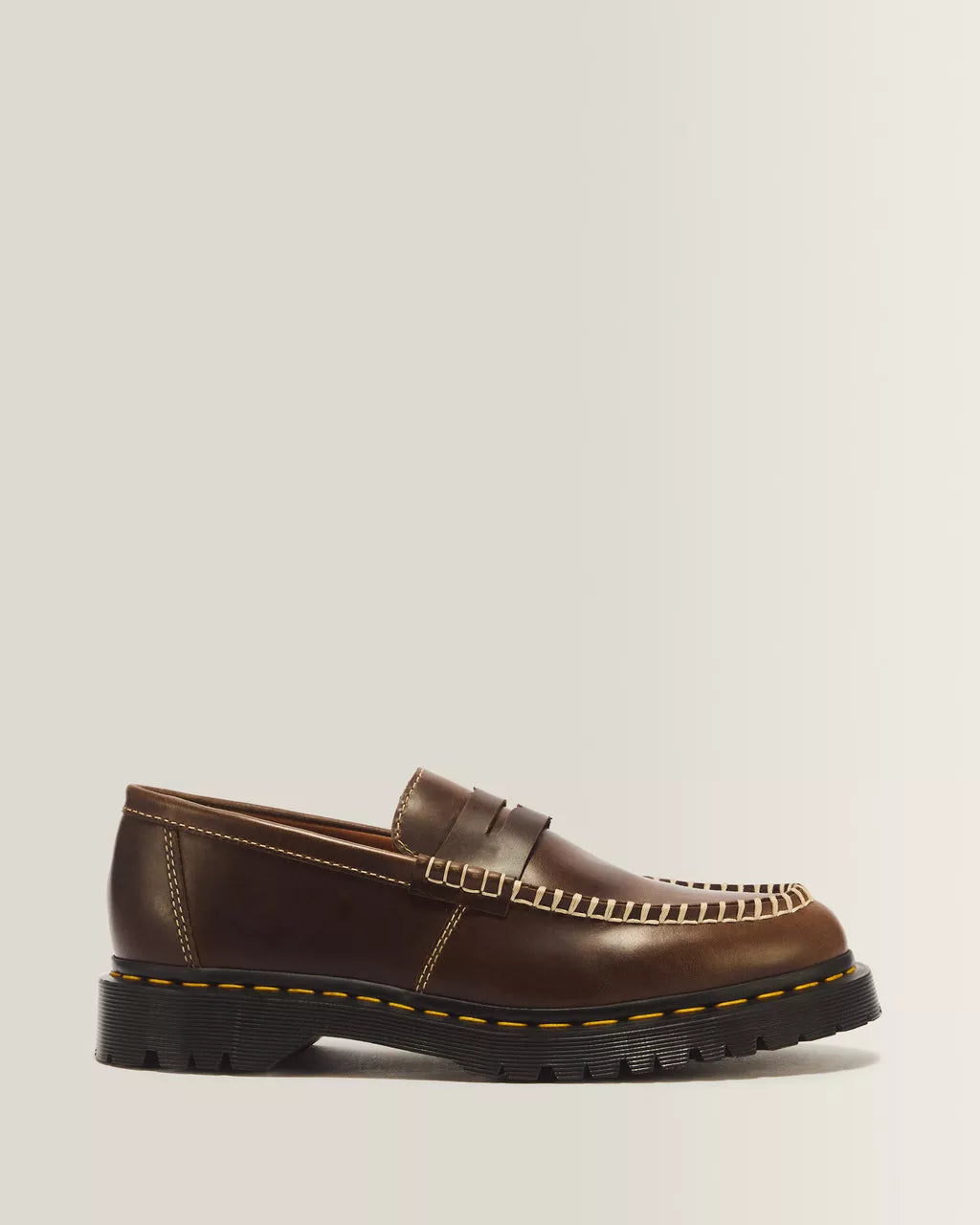 A single brown leather penny loafer with a chunky black sole, featuring decorative stitching and contrast yellow stitching along the edge, shown against a light gray background.