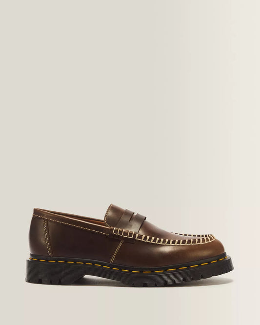A single brown leather penny loafer with a chunky black sole, featuring decorative stitching and contrast yellow stitching along the edge, shown against a light gray background.