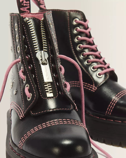 A close-up of a black leather combat boot with pink contrast stitching and pink laces, featuring a prominent silver zipper on the side. A second matching boot is partially visible in the background against a light neutral backdrop.