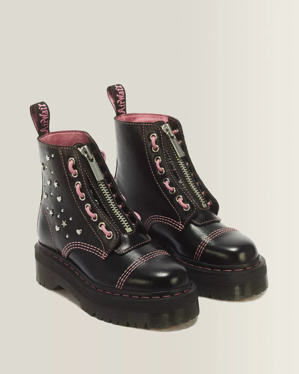 A pair of black leather ankle boots with pink laces, pink stitching, silver zippers, and heart-shaped studs, shown against a light gray background.