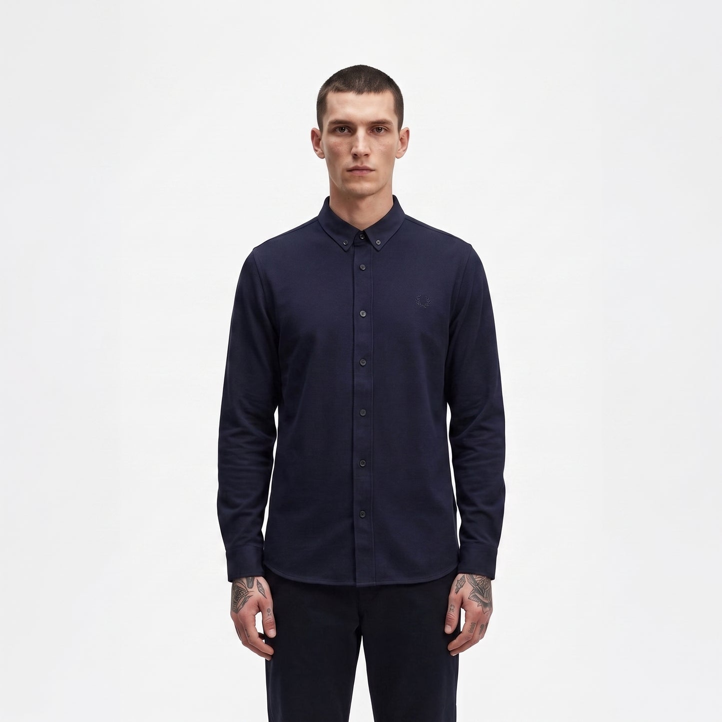 A man wearing a dark navy blue long-sleeve button-down shirt and matching dark trousers, standing against a plain white studio background.