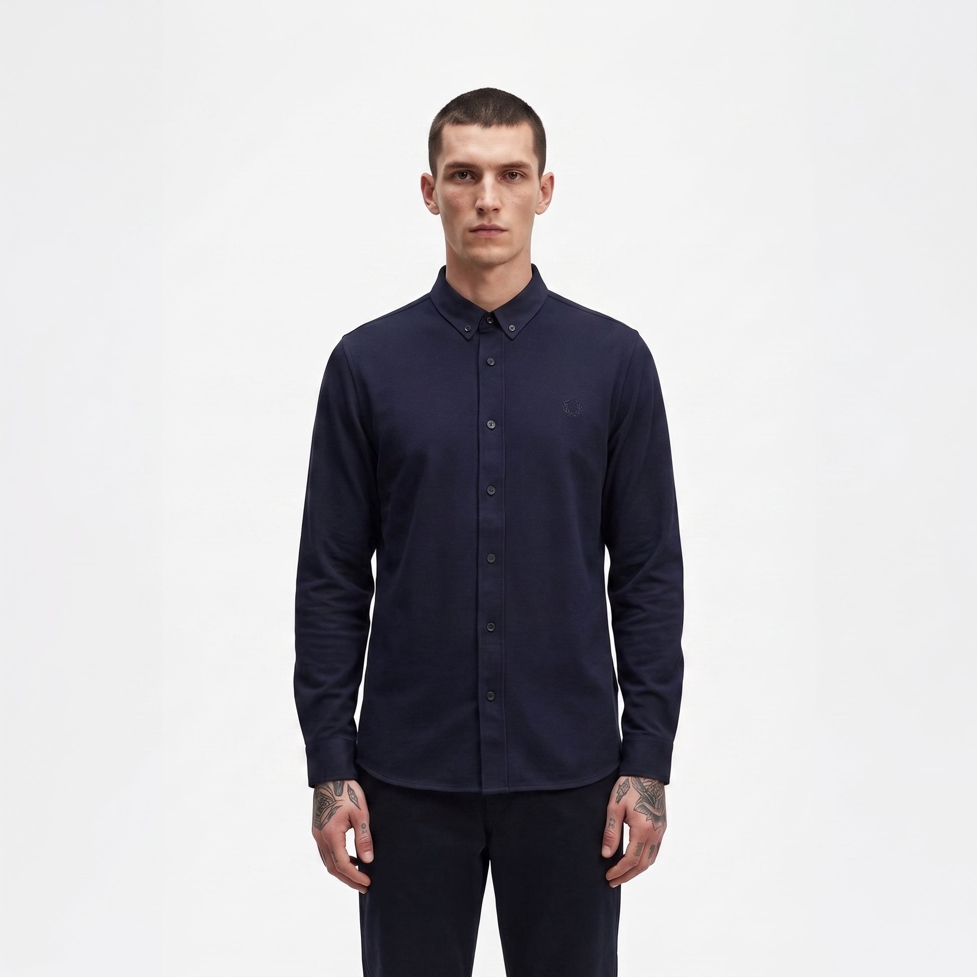 A man wearing a dark navy blue long-sleeve button-down shirt and matching dark trousers, standing against a plain white studio background.