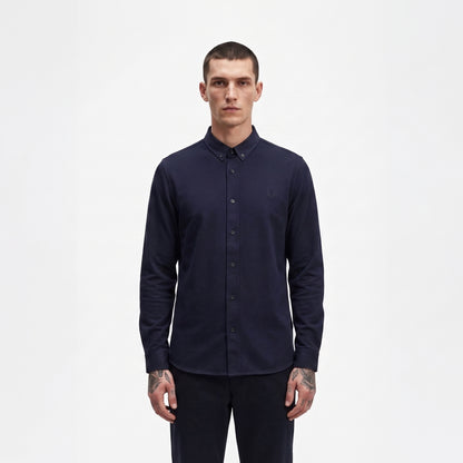 A man wearing a dark navy blue long-sleeve button-down shirt and matching dark trousers, standing against a plain white studio background.