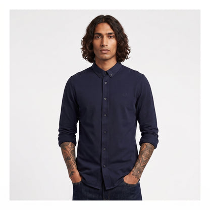 A man with long dark hair and tattoos wearing a dark navy long-sleeve button-down shirt, standing against a light gray background.