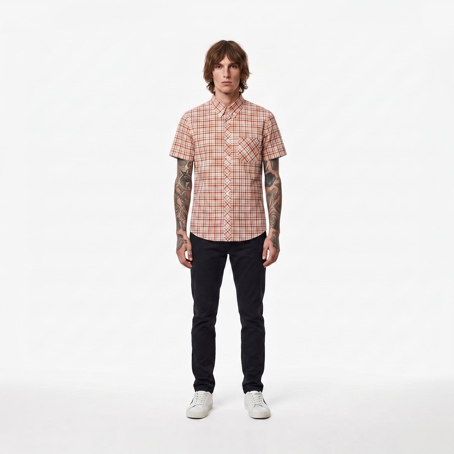 A man stands against a plain white studio background wearing a short-sleeve button-up shirt with a red and beige plaid pattern, dark pants, and white sneakers.