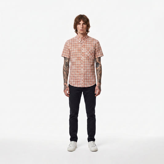 A man stands against a plain white studio background wearing a short-sleeve button-up shirt with a red and beige plaid pattern, dark pants, and white sneakers.