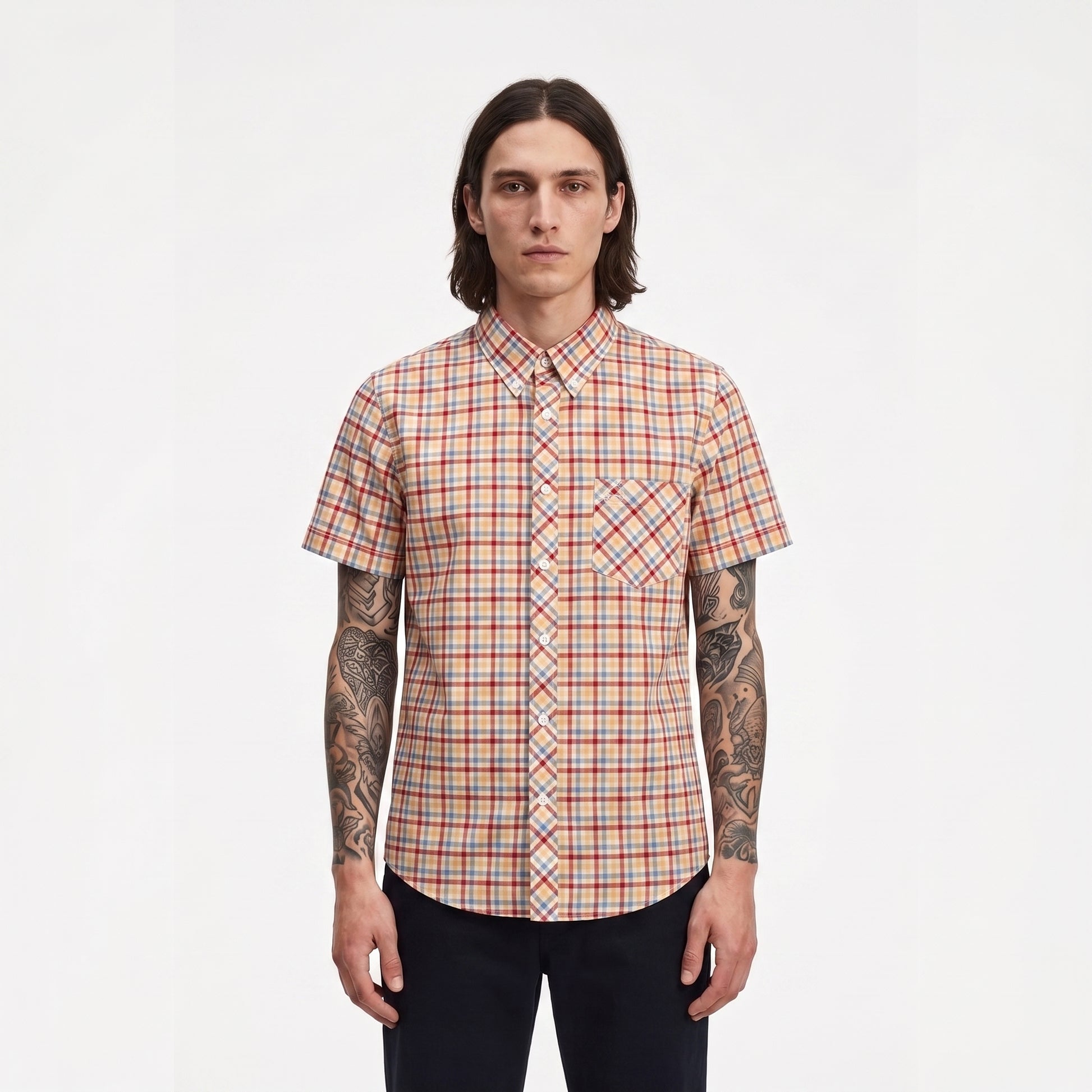 A man with long dark hair and tattoos stands against a plain white background wearing a short-sleeve button-up shirt with a red, orange, and blue plaid pattern.