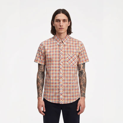 A man with long dark hair and tattoos stands against a plain white background wearing a short-sleeve button-up shirt with a red, orange, and blue plaid pattern.