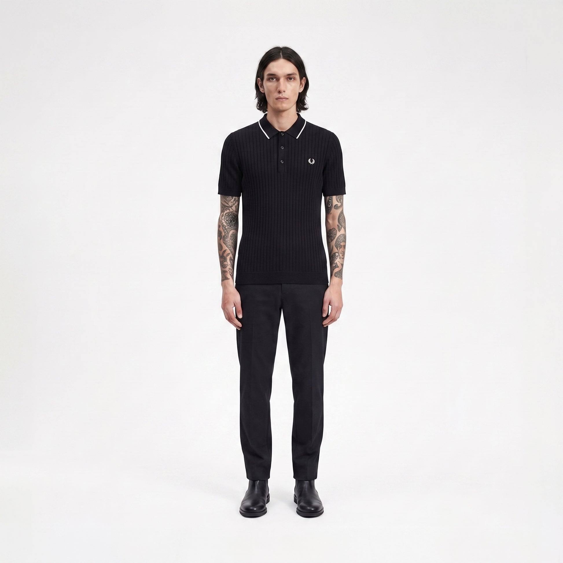 A man stands facing forward in a black ribbed short-sleeve polo shirt with white piping and a small embroidered laurel wreath logo on the chest, paired with black trousers and black shoes, against a plain light gray studio background.