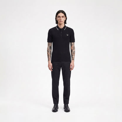 A man stands facing forward in a black ribbed short-sleeve polo shirt with white piping and a small embroidered laurel wreath logo on the chest, paired with black trousers and black shoes, against a plain light gray studio background.