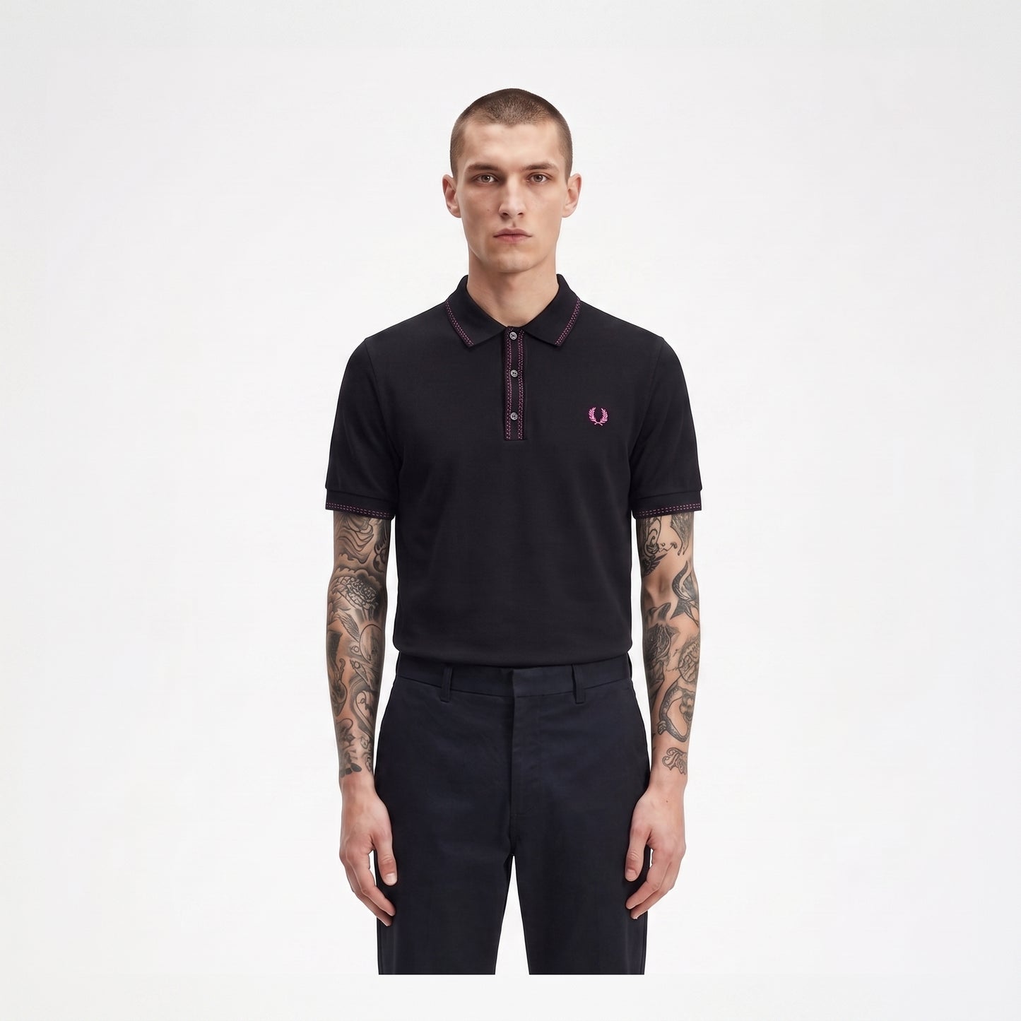 A man modeling a black short-sleeve polo shirt with a dark red collar and placket, featuring the signature pink and white embroidered laurel branch logo on the left chest, paired with dark trousers against a plain white background.