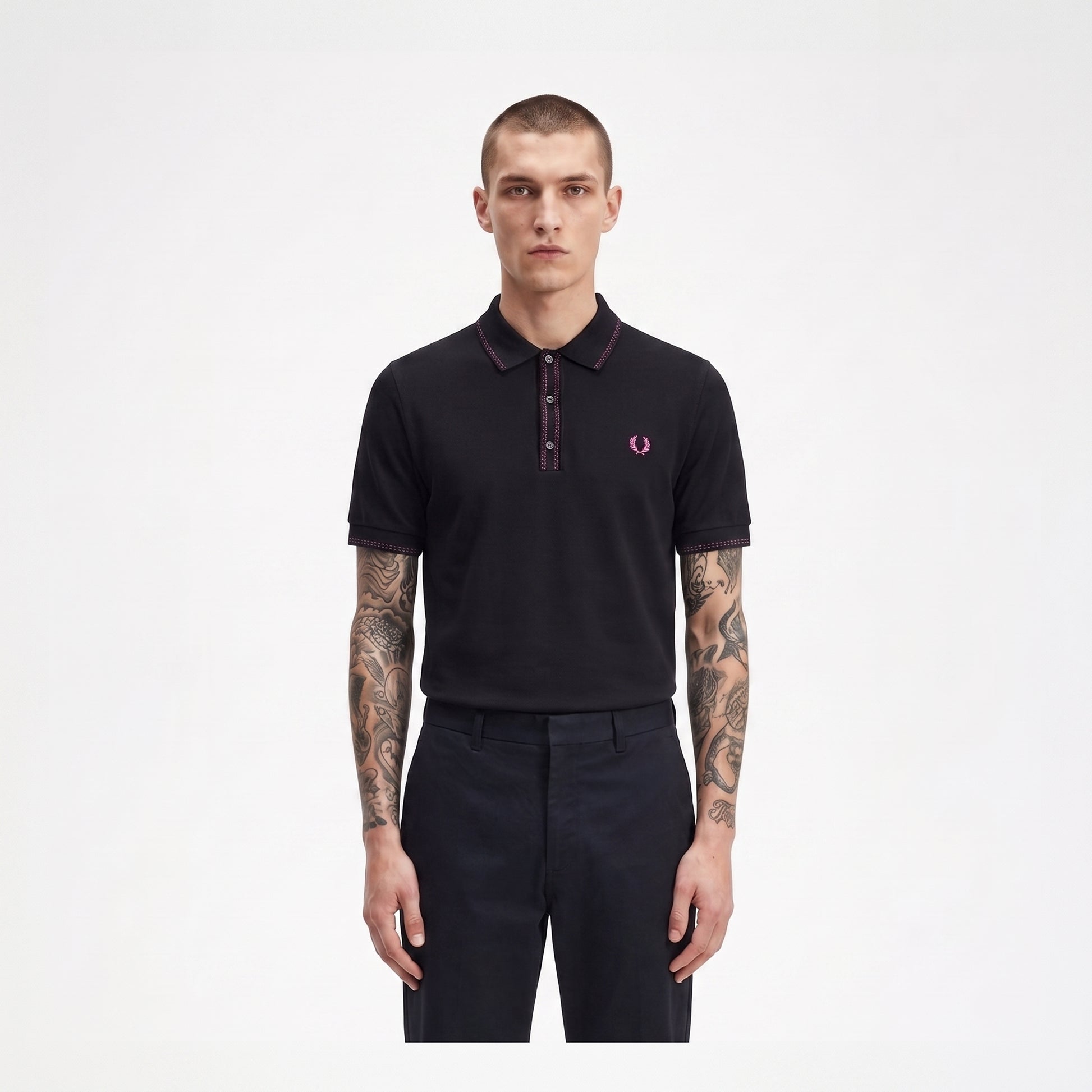 A man modeling a black short-sleeve polo shirt with a dark red collar and placket, featuring the signature pink and white embroidered laurel branch logo on the left chest, paired with dark trousers against a plain white background.