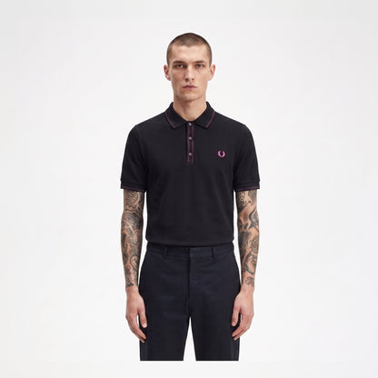 A man modeling a black short-sleeve polo shirt with a dark red collar and placket, featuring the signature pink and white embroidered laurel branch logo on the left chest, paired with dark trousers against a plain white background.