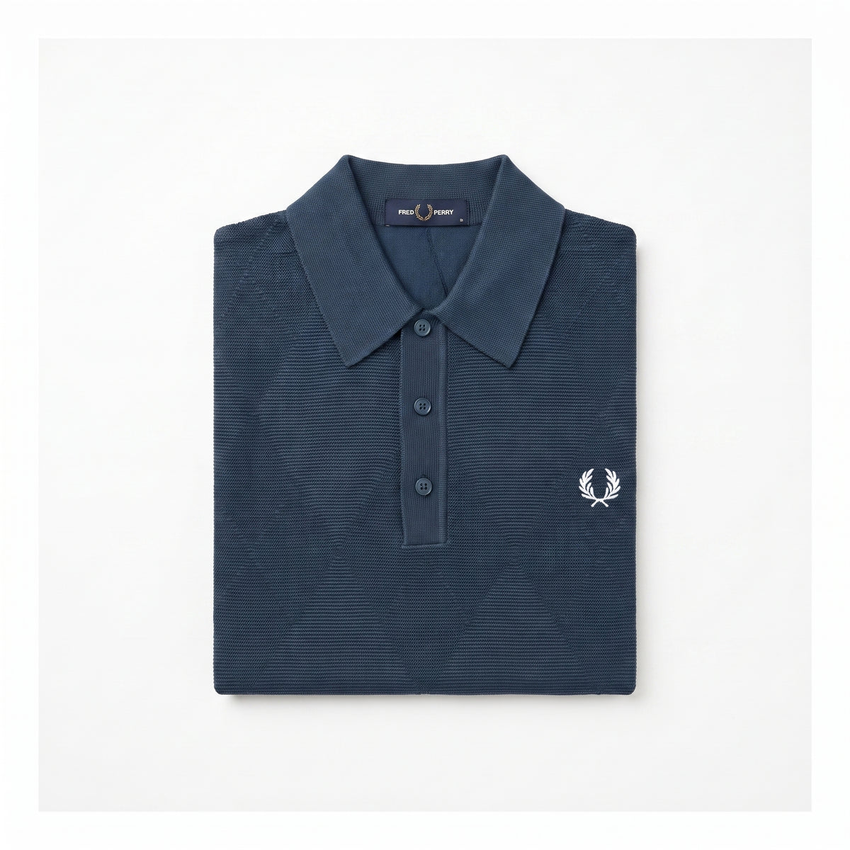A folded navy blue polo shirt with a textured waffle knit pattern, featuring a white embroidered laurel wreath logo on the left chest, set against a plain white background.