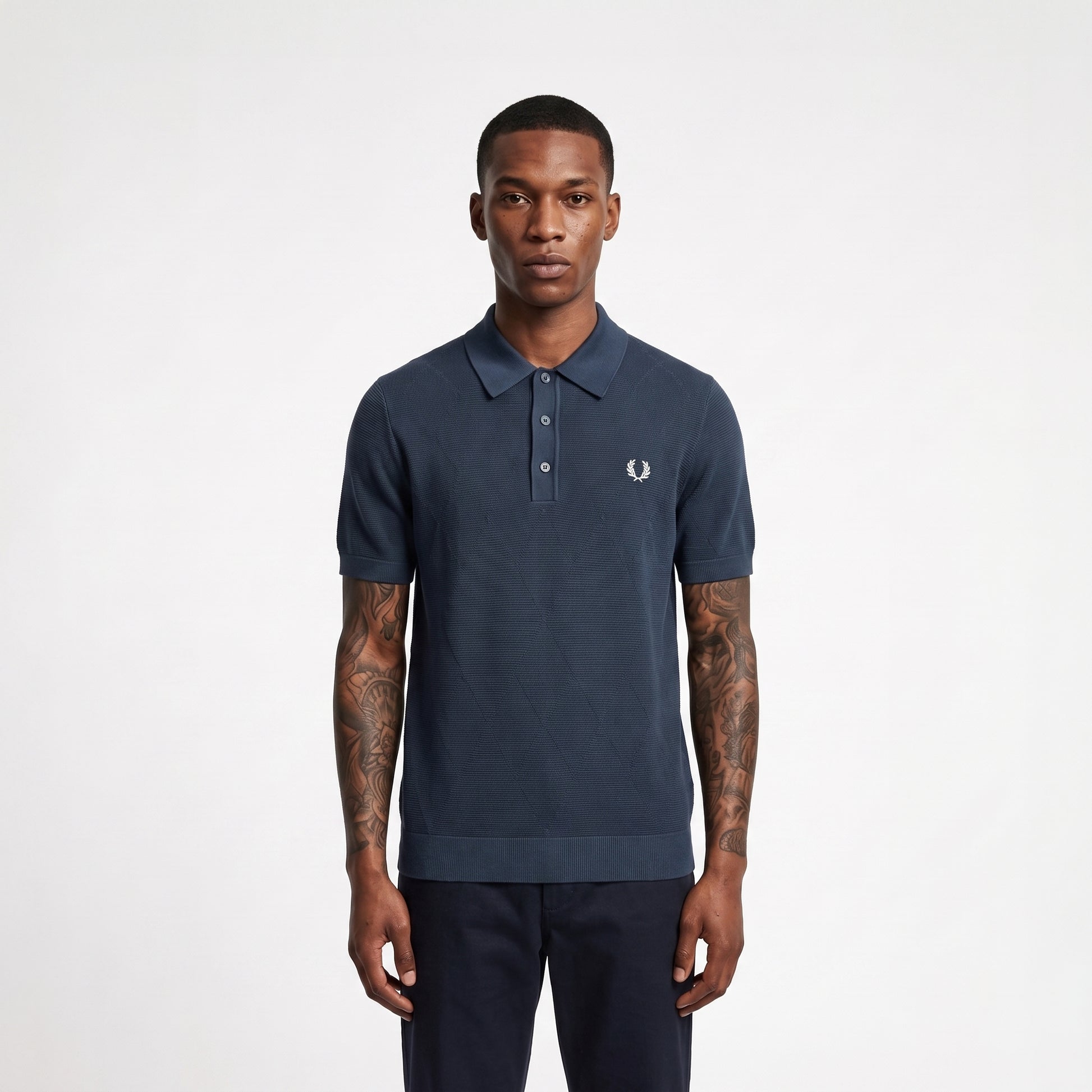 A man wearing a navy blue short-sleeve polo shirt with a small white embroidered logo on the chest, paired with dark trousers, standing against a light gray studio background.
