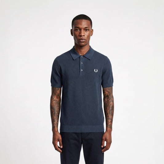 A man wearing a navy blue short-sleeve polo shirt with a small white embroidered logo on the chest, paired with dark trousers, standing against a light gray studio background.