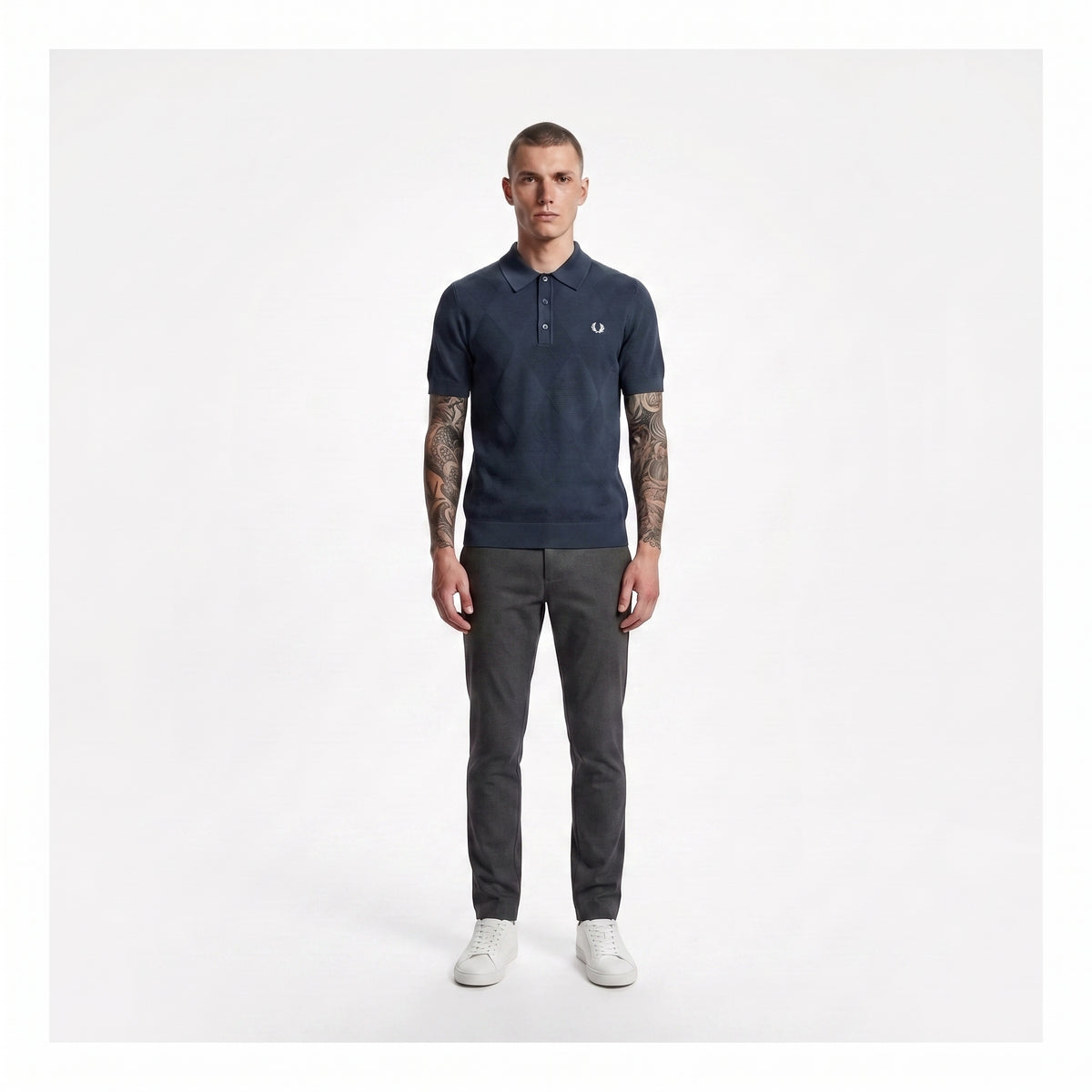 A man modeling a navy blue polo shirt with a small embroidered logo on the chest, paired with dark gray trousers and white sneakers, standing against a plain light gray background.