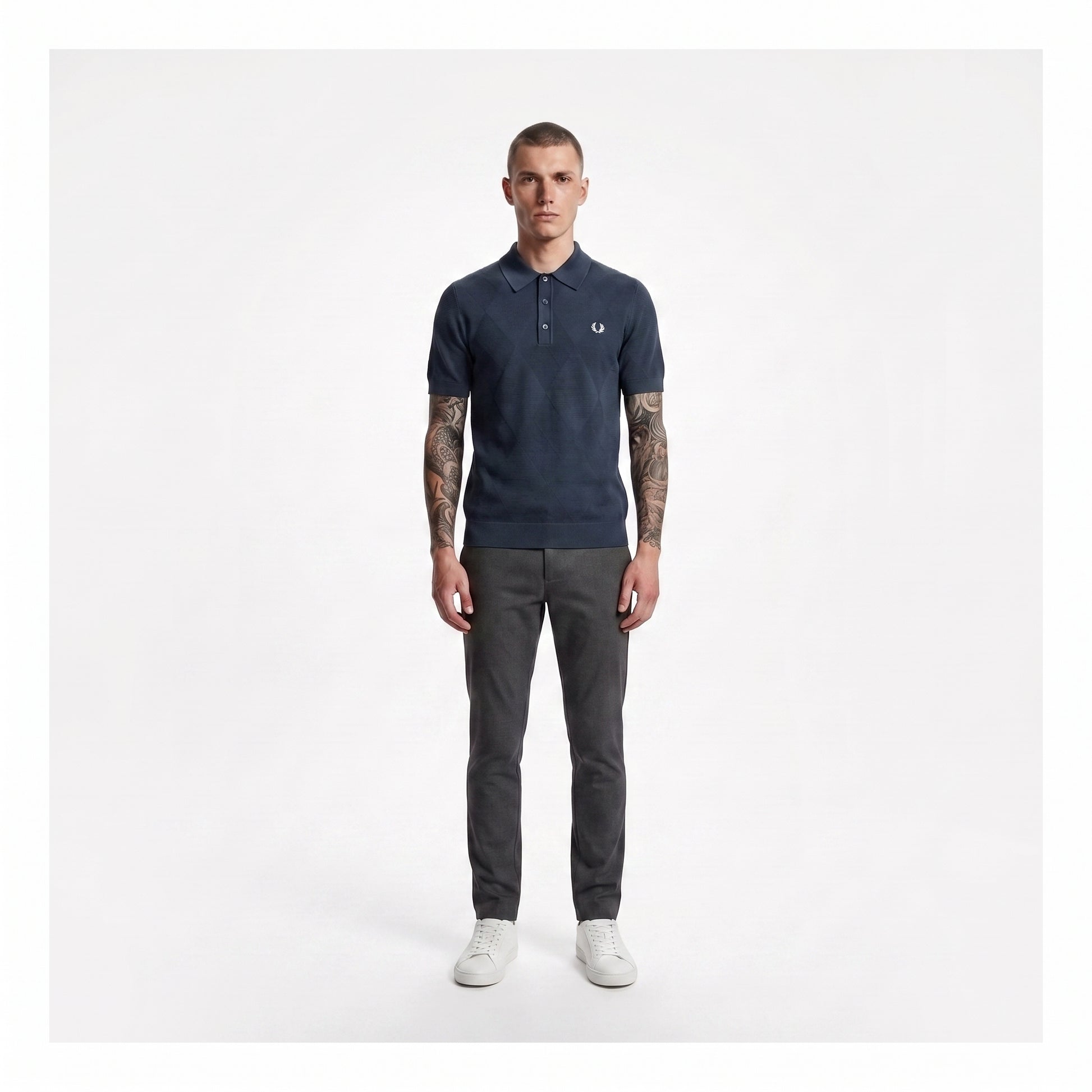 A man modeling a navy blue polo shirt with a small embroidered logo on the chest, paired with dark gray trousers and white sneakers, standing against a plain light gray background.