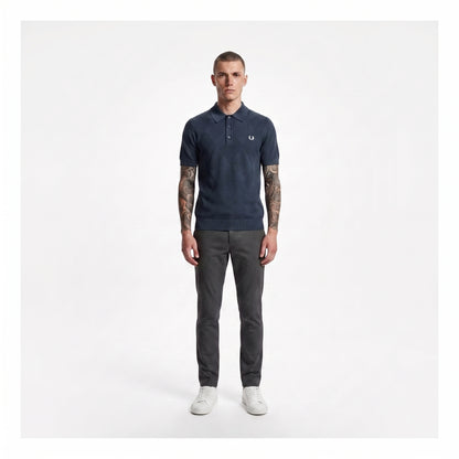 A man modeling a navy blue polo shirt with a small embroidered logo on the chest, paired with dark gray trousers and white sneakers, standing against a plain light gray background.