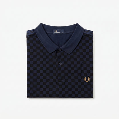 A folded navy blue polo shirt with a dark blue and black checkerboard pattern, featuring a small gold embroidered laurel wreath logo on the left chest. The brand label inside the collar reads ‘Fred Perry’