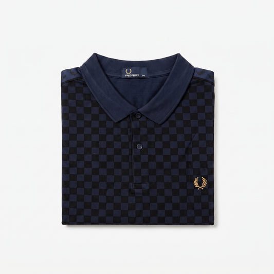 A folded navy blue polo shirt with a dark blue and black checkerboard pattern, featuring a small gold embroidered laurel wreath logo on the left chest. The brand label inside the collar reads ‘Fred Perry’