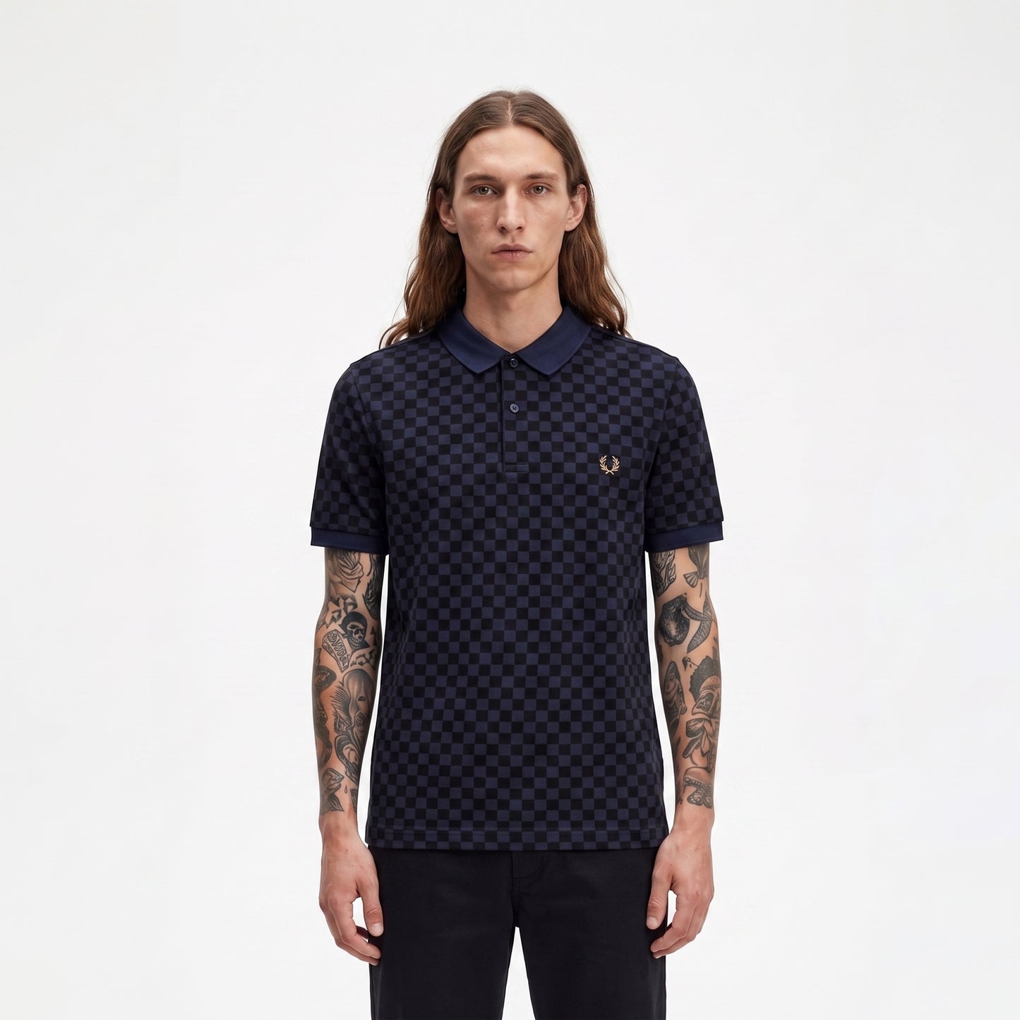 A man with long brown hair and tattooed arms wearing a dark navy blue polo shirt with a small embroidered laurel wreath logo on the chest, standing against a plain white background.
