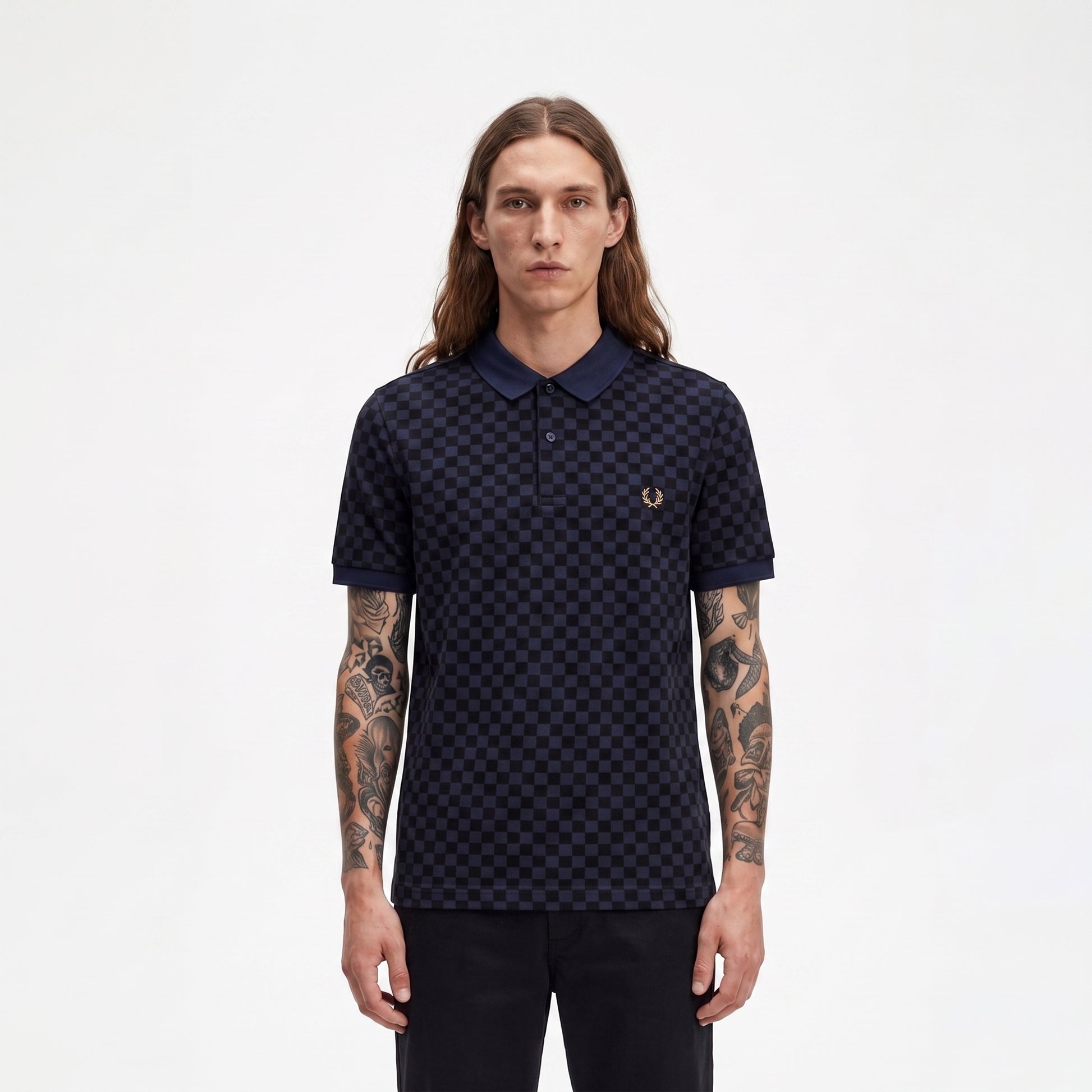 A man with long brown hair and tattooed arms wearing a dark navy blue polo shirt with a small embroidered laurel wreath logo on the chest, standing against a plain white background.