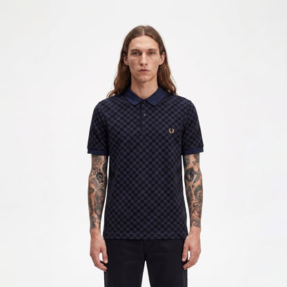 A man with long brown hair and tattooed arms wearing a dark navy blue polo shirt with a small embroidered laurel wreath logo on the chest, standing against a plain white background.