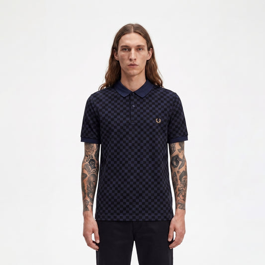 A man with long brown hair and tattooed arms wearing a dark navy blue polo shirt with a small embroidered laurel wreath logo on the chest, standing against a plain white background.