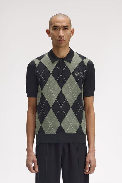 Man wearing a black and green argyle patterned sweater vest over a black polo shirt on a white background