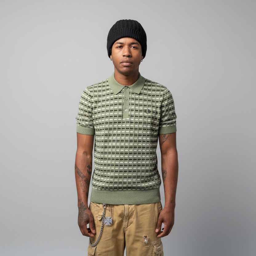 A person wearing a Fred Perry boucle ribbed knitted shirt in olive mint color, paired with a beanie and shorts, standing in front of a wall with graffiti.